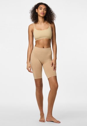 Femme debout pieds nus portant un crop top beige sans manches et un short cycliste taille haute ajusté sur fond uni.