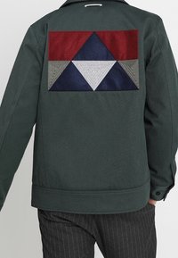 Grüne Jacke mit geometrischem Patchwork-Design in Rot, Marineblau, Grau und Weiß auf dem Rücken. Strukturierter Stoff mit Kragen und Knopfmanschetten.
