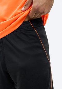 Χέρι που σηκώνει ένα πορτοκαλί αθλητικό μπλουζάκι, αποκαλύπτοντας μαύρο αθλητικό σορτς με πορτοκαλί λεπτομέρειες πάνω σε απλό φόντο.