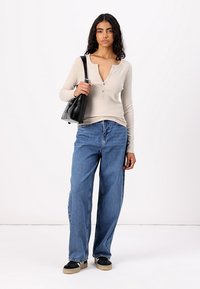 Top bej cu mânecă lungă, cu model ribbed și guler cu nasturi, purtat cu blugi denim bleumarin largi și adidași negri, ținând în mână o geantă neagră.