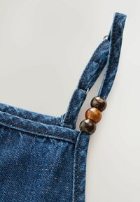 Sangle de vêtement en denim avec trois perles rondes brillantes en marron foncé et ambre attachées près d'un anneau métallique sur fond clair.