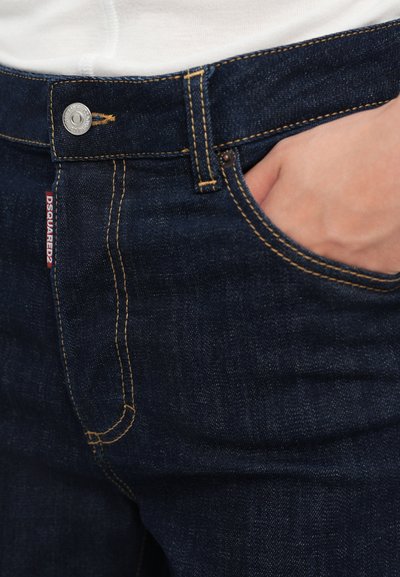 Jeanși din denim bleu închis cu cusături aurii, având un buzunar frontal, închidere cu buton metalic și o etichetă de marcă vizibilă pe lateral.
