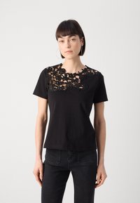 Sort bomulds t-shirt med en blonderdesign over halsudskæringen. Korte ærmer og klassisk pasform. Bæres med mørke jeans, ensfarvet baggrund.