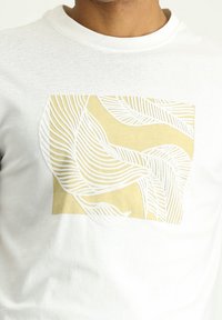 Camiseta blanca con un gráfico de cuadrado amarillo que presenta intrincados patrones de hojas en línea blanca. Material de algodón suave y diseño de cuello redondo.