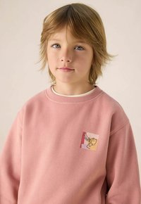 Roze katoenen sweatshirt met een kleine meerkleurige grafische patch van een karakter dat op de borst een trompet speelt. Zachte textuur.