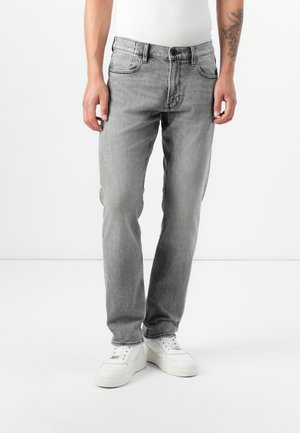 En ung mann står iført grå jeans og en tettsittende hvit tanktop, med hvite sneakers, mot en ensfarget hvit bakgrunn.