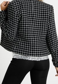 Czarnoskalisty żakiet w biało-czarną kratę o teksturowanym materiale. Posiada prosty krój i lekki połysk. Połączony z topem wyszywanym cekinami.