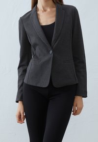 Blazer gris hecho de tela lisa, con cierre de un solo botón, solapas con muesca y dos bolsillos delanteros. Estilizado sobre una blusa negra.