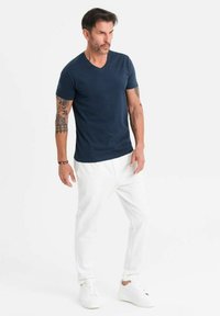 T-shirt a V blu navy, abbinato a pantaloni bianchi tapered e sneaker bianche. Il modello ha tatuaggi visibili sulle braccia, in posa su uno sfondo neutro.