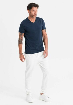 T-shirt bleu marine en v, associé à un pantalon blanc ajusté et des baskets blanches. Le mannequin a des tatouages visibles sur les bras et pose sur un fond neutre.
