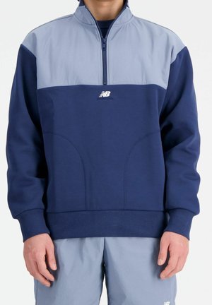 Mężczyzna ubrany w granatową i jasnoniebieską bluzę z suwakiem do ćwiartki, z małym białym logo New Balance na środku klatki piersiowej.