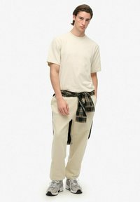 Camiseta beige de manga corta con cuello redondo, combinada con pantalones beige holgados y una camisa de franela a cuadros atada a la cintura, con líneas en verde oscuro y amarillo.