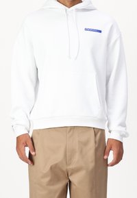 Sudadera blanca con un bolsillo frontal, cordones y logo bordado azul. Tejido suave, corte relajado y puños acanalados en las muñecas y la cintura.