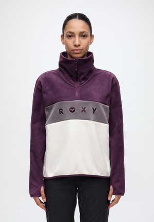 Une femme porte un pull polaire ROXY violet, gris et blanc, avec un col montant zippé et un pantalon noir, sur un fond uni.
