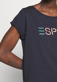 Camiseta de algodón azul marino con mangas cortas enrolladas y la palabra "ESP" en letras verdes, rosas y amarillas en la parte delantera.