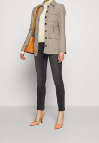 Femme portant un col roulé beige, une veste à carreaux, un jean skinny foncé, des escarpins pêche et un sac à bandoulière en cuir tan.