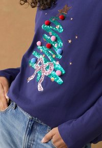 Marineblå langærmet skjorte med et palietbelagt juletre-design, farverige pomponer, en lyserød sløjfe og stjernedetaljer. Denimjeans er synlige.