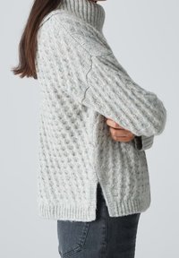 Personne aux longs cheveux bruns portant un pull à col roulé en maille torsadée gris clair et un jean foncé, les bras croisés, vue de côté.