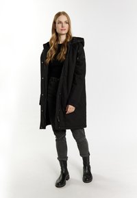 Sort isoleret parka med hætte, trykknap- og lynlåslukning, med lange ærmer og sidelommer, båret over mørke jeans og sorte støvler.