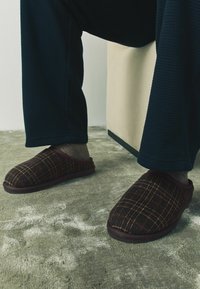 Braune karierte Hausschuhe aus weichem Stoff mit einem tief ausgeschnittenen Design, kombiniert mit dunkel strukturierten Schlafanzughosen auf einem strukturierten grauen Boden.