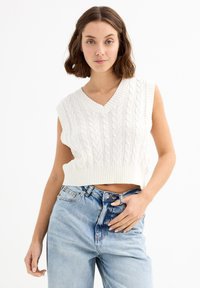 Mujer joven con cabello castaño ondulado que lleva un suéter sin mangas de punto blanco y jeans de tiro alto de color azul claro, sobre un fondo blanco.