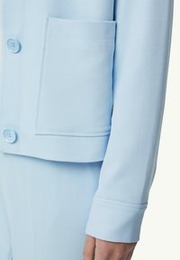 Jasnoniebieska marynarka z fakturowanej tkaniny, z jedną kieszenią i detalem w postaci guzików. Rękawy są dopasowane, co tworzy zarysowane sylwetkę.
