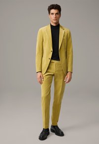 Geel corduroy pak met een op maat gemaakte pasvorm, een enkelzijdig blazer en slanke broek, gecombineerd met een zwarte coltrui en zwarte schoenen.