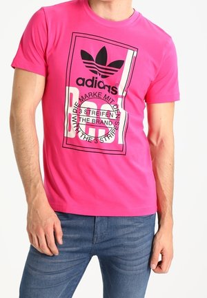 Mann trägt ein leuchtend pinkes Adidas-T-Shirt mit schwarzem Logo und Schriftzug, kombiniert mit blauen Jeans, steht vor weißem Hintergrund.