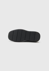 Black rubber shoe sole with a textured surface, featuring horizontal grooves for traction and logo detail. Smooth edges and round shape.