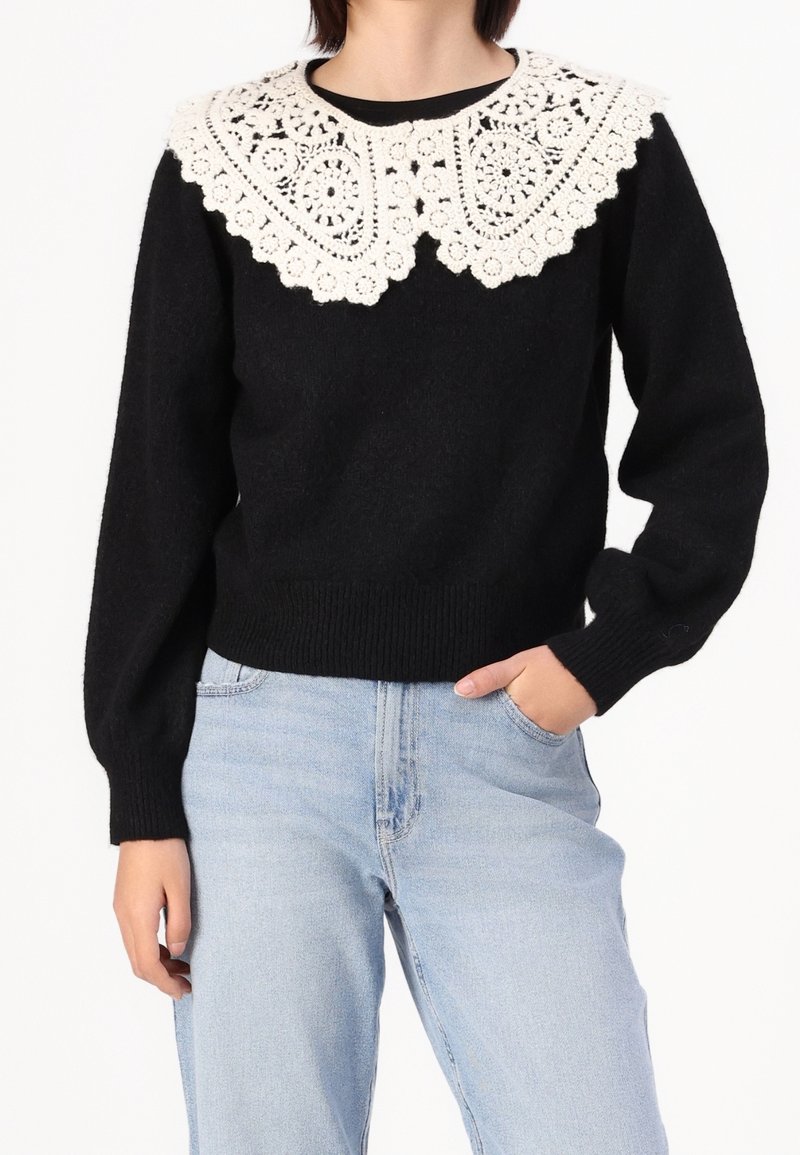 Pull noir avec des manches volumineuses et un large col en dentelle blanche. Le matériau semble doux, avec un ourlet et des poignets côtelés.