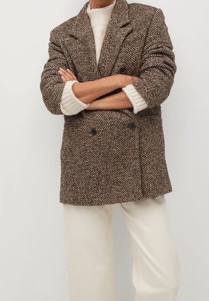 Personne portant un blazer croisé en chevrons marron sur un pull en maille blanche à manches retroussées et un pantalon large crème.