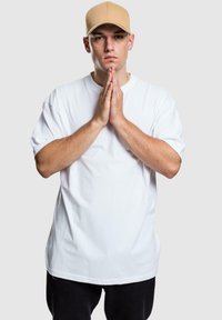 Witte katoenen t-shirt met korte mouwen, gecombineerd met een lichtbruine baseballcap. Model staat met de handen voor elkaar.