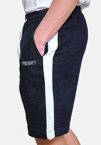 Shorts in denim scuro con strisce laterali bianche, vita elastica e tasche laterali. Presentano il logo di Armani Exchange sulla tasca sinistra.