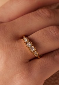 Gouden ring met vijf ronde, heldere stenen in ingewikkelde, gestructureerde settings, gedragen op een hand met een gladde huidstructuur.