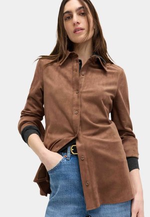 Bruin suède overhemd met knopen aan de voorkant, spitse kraag en opgerolde mouwen met zwarte manchetagaccenten, gecombineerd met blauwe spijkerbroek.