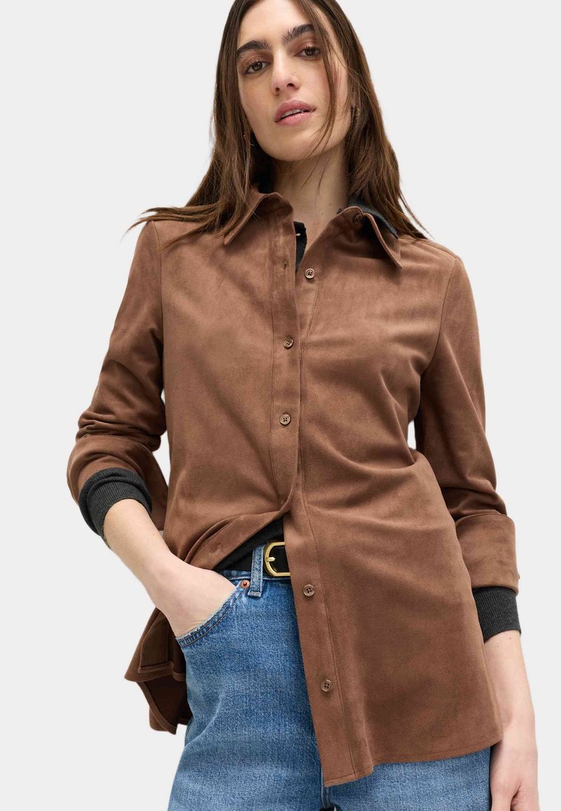 Camicia marrone in suede con chiusura frontale a bottoni, colletto appuntito e maniche arrotolate con dettagli neri sui polsini, abbinata a jeans di denim blu.