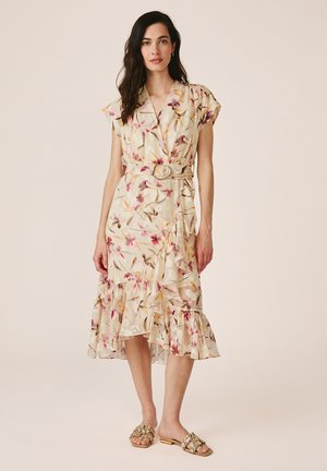 Donna con vestito a portafoglio beige a fiori, con cintura, orlo arricciato e maniche corte, abbinato a sandali bassi oro decorati.