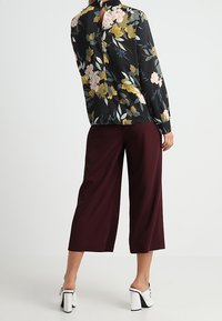 Kwiatowa czarna bluzka z długimi rękawami i wycięciem z tyłu, zestawiona z bordowymi spodniami o szerokich nogawkach oraz białymi klamerkowymi mule.