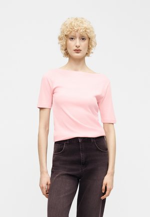 Mujer con cabello corto y rizado rubio, que lleva una camiseta de manga corta de color rosa claro y jeans oscuros de cintura alta, de pie contra un fondo claro y liso.