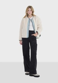 Chaqueta de forro polar crema con detalles en azul claro, botones a presión y dos bolsillos, combinada con pantalones de pana negros y botas de tobillo grises.