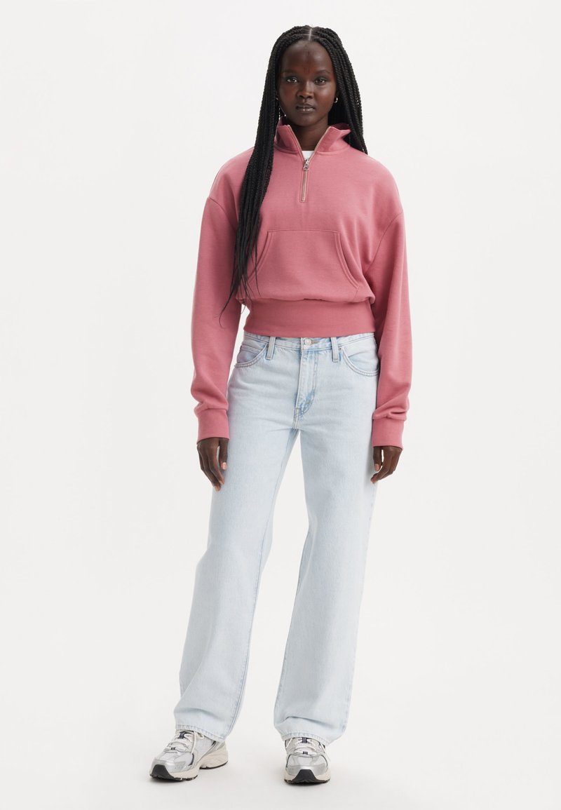 Felpa con zip di un rosa polveroso e tasca frontale, abbinata a jeans a gamba dritta blu chiaro e sneaker grigie, su sfondo bianco.