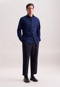 Camicia di tessuto blu navy con maniche lunghe e chiusura frontale con bottoni, abbinata a pantaloni scuri su misura e scarpe di pelle nere.