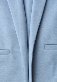 Blazer bleu clair avec revers crantés et deux poches passepoilées horizontales sur le devant, montrant la texture du tissu et les détails des coutures.