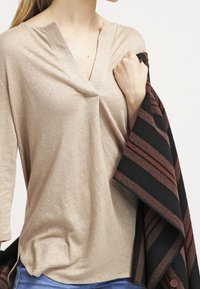 Top beige con texture, con scollatura a V e maniche a tre quarti, abbinato a un wrap a strisce nere e marroni. Il tessuto sembra leggero.