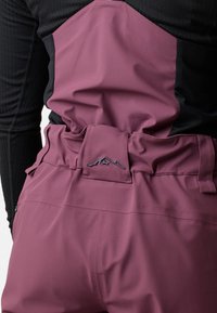 Vue arrière d'une personne portant un pantalon mauve taille haute avec une ceinture ajustable et un haut noir à manches longues, mettant en avant le logo montagne du pantalon.