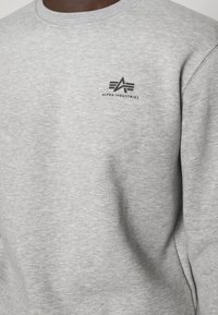 Felpa grigia in tessuto morbido, con scollo rotondo e un piccolo logo nero con tre linee stilizzate sul petto.