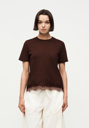 Γυναίκα με κοντά σκούρα μαλλιά που φορά καφέ κοντομάνικη μπλουζα με δαντέλα και άνετο λευκό παντελόνι, στέκεται μπροστά σε απλό φόντο.