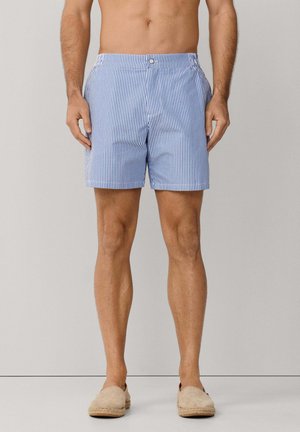 Mężczyzna w niebiesko-białych, pionowo paskowanych szortach i beżowych butach wsuwanych, stojący na tle jednolitego szarego tła.