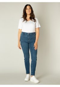Witte V-hals shirt, blauwe hoge skinny jeans en witte sneakers. De jeans hebben een knoop aan de voorkant en riemlussen, met een gladde textuur.