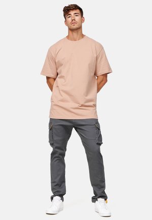 Młody mężczyzna stoi z rękami za plecami, ubrany w beżowy oversize'owy T-shirt, szare spodnie cargo i białe trampki na jednolitym tle.